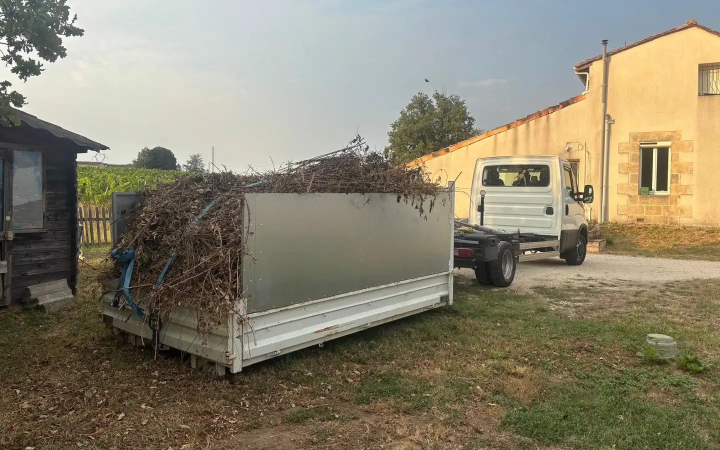 Camion à bras amplirol et benne remplie de branchages dans une cour, devant une maison